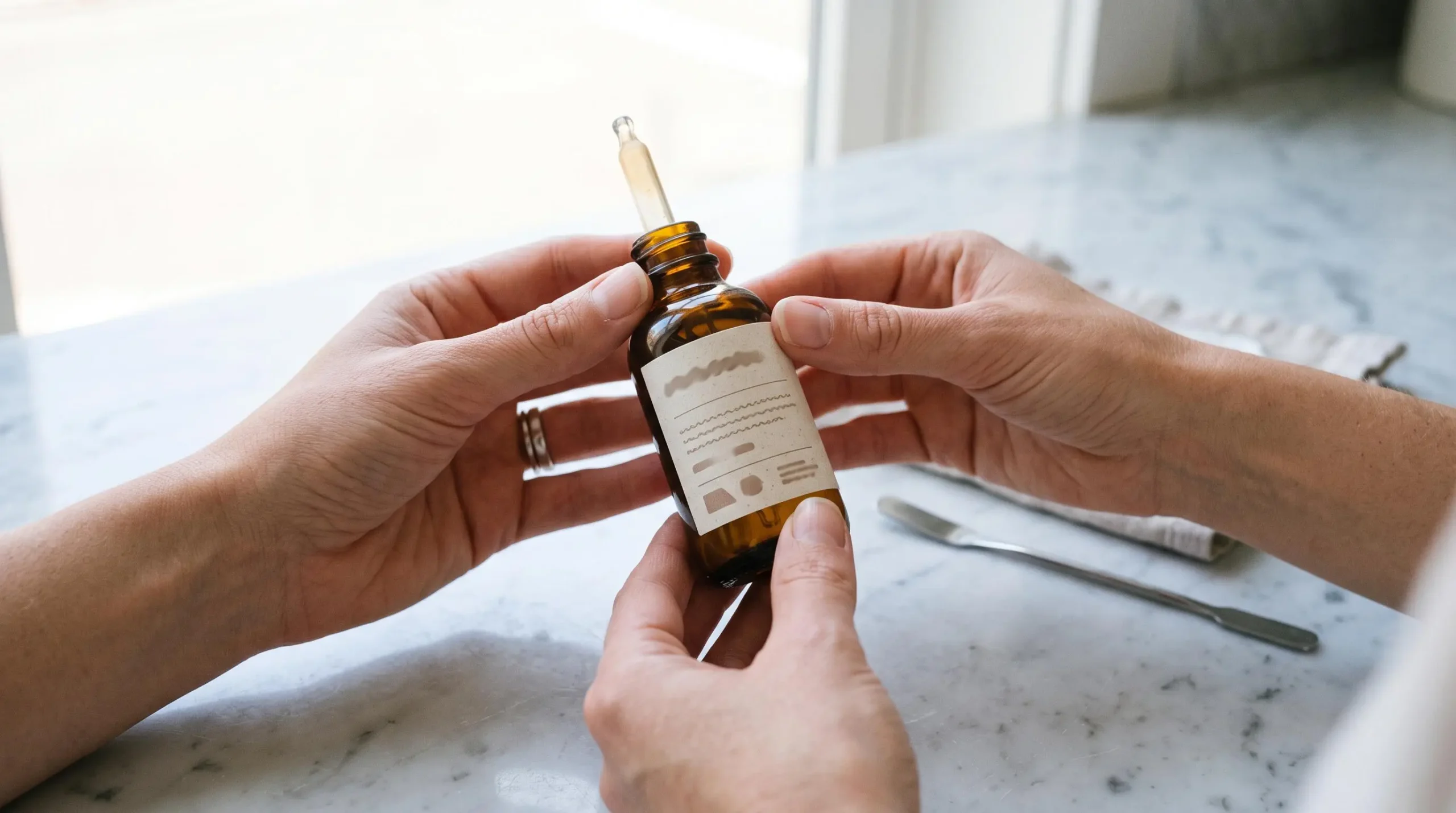 Mains de femme examinant l'étiquette d'un flacon d'huile de CBD sur une surface claire